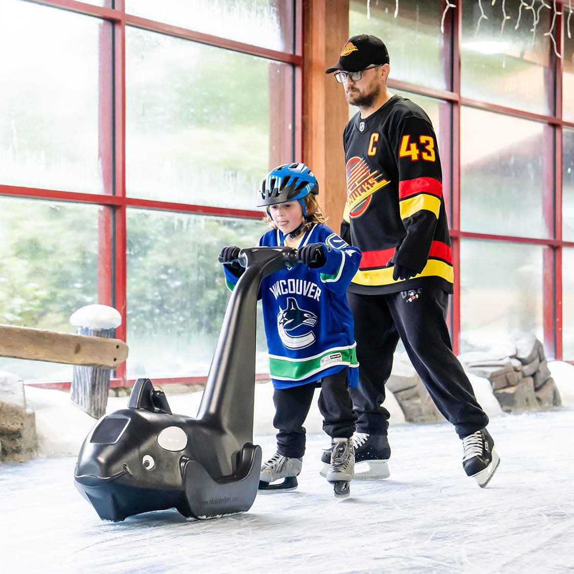 Young skater pushing a Black and White Orca Skate Helper on an indoor rink with a parent skating behind